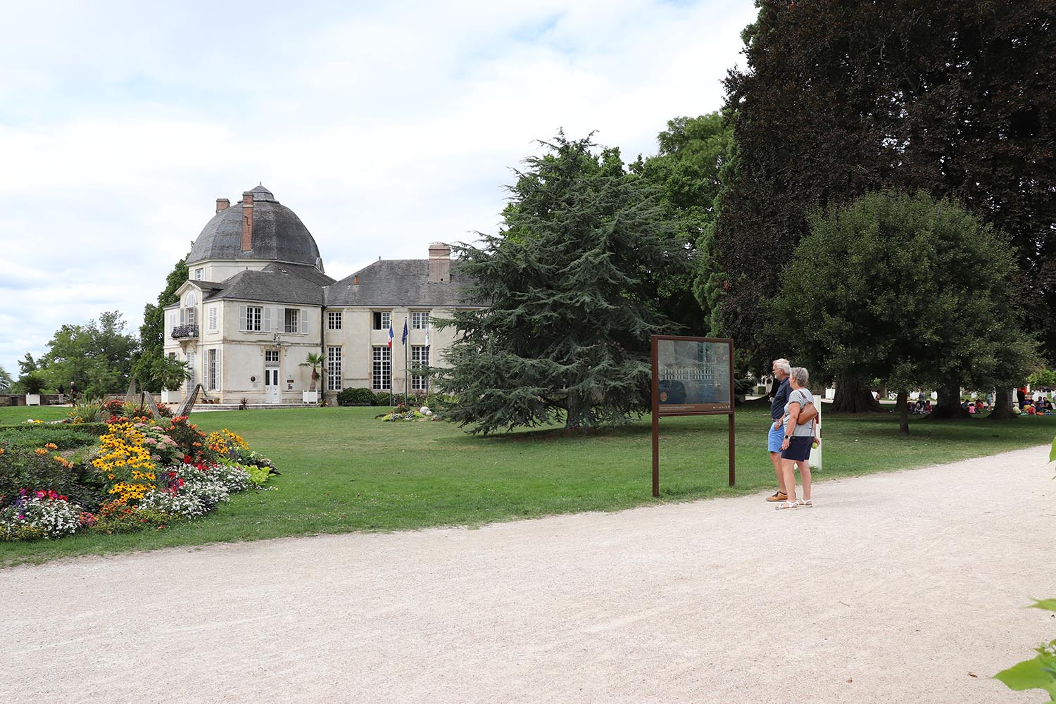 Château historique en arrière-plan avec dispositif de médiation patrimoniale transparent observé par des visiteurs dans un parcours aménagé