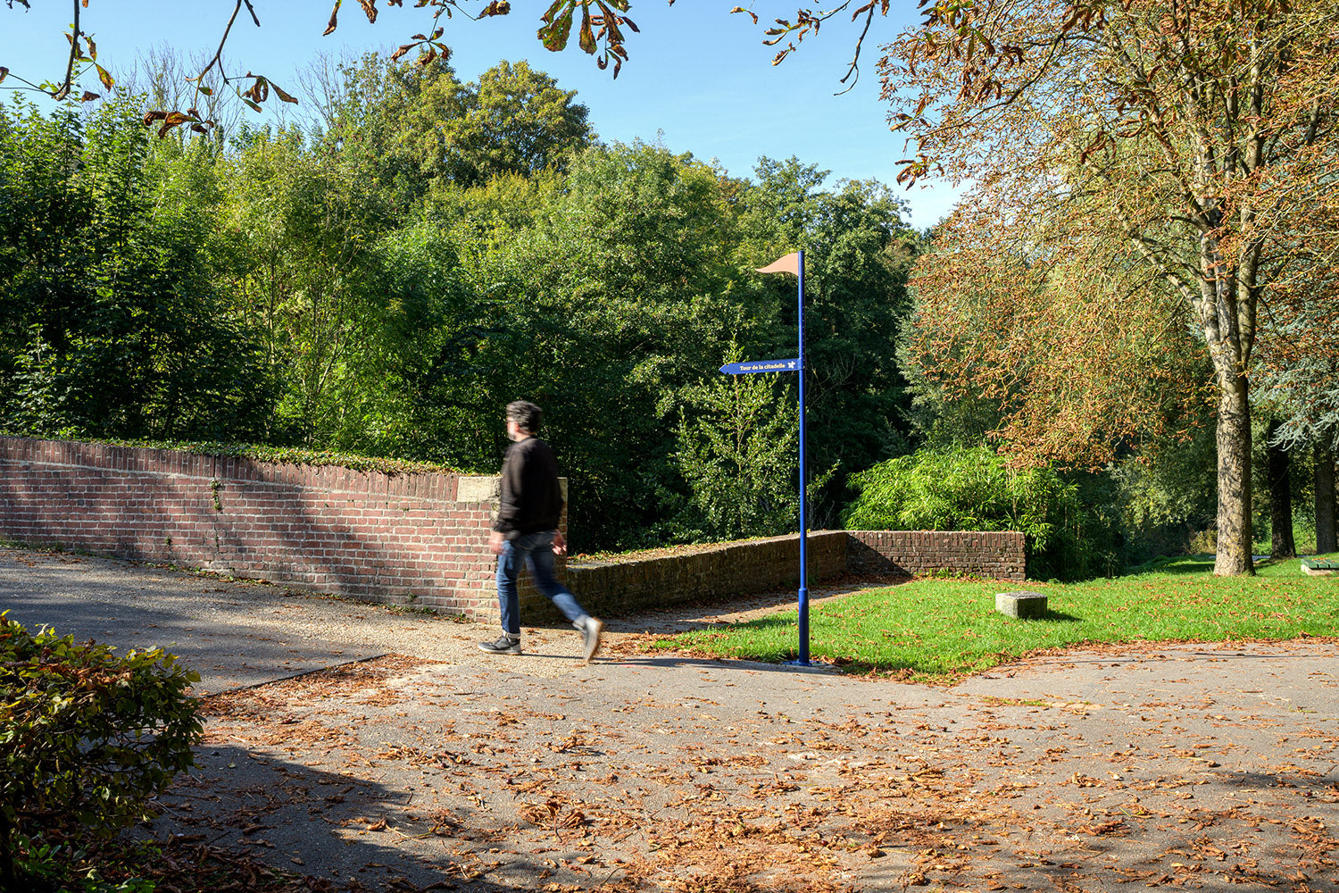 Piéton en promenade sur un parcours mi-urbain mi-naturel avec mât de signalétique identitaire pour la mobilité douce