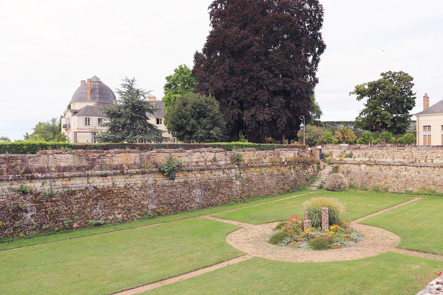 Vue éloignée du château depuis un espace public avec vestiges des murs anciens et jardin géométrique mettant en valeur le patrimoine